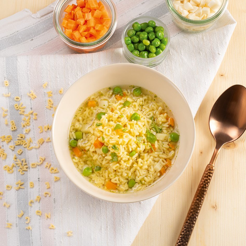 [Translate to Englisch:] Schale mit Buchstabennudelsuppe mit Erbsen und Karotten, daneben drei Gläser mit gewürfelten Karotten, Erbsen und Sellerie, brauner Löffel