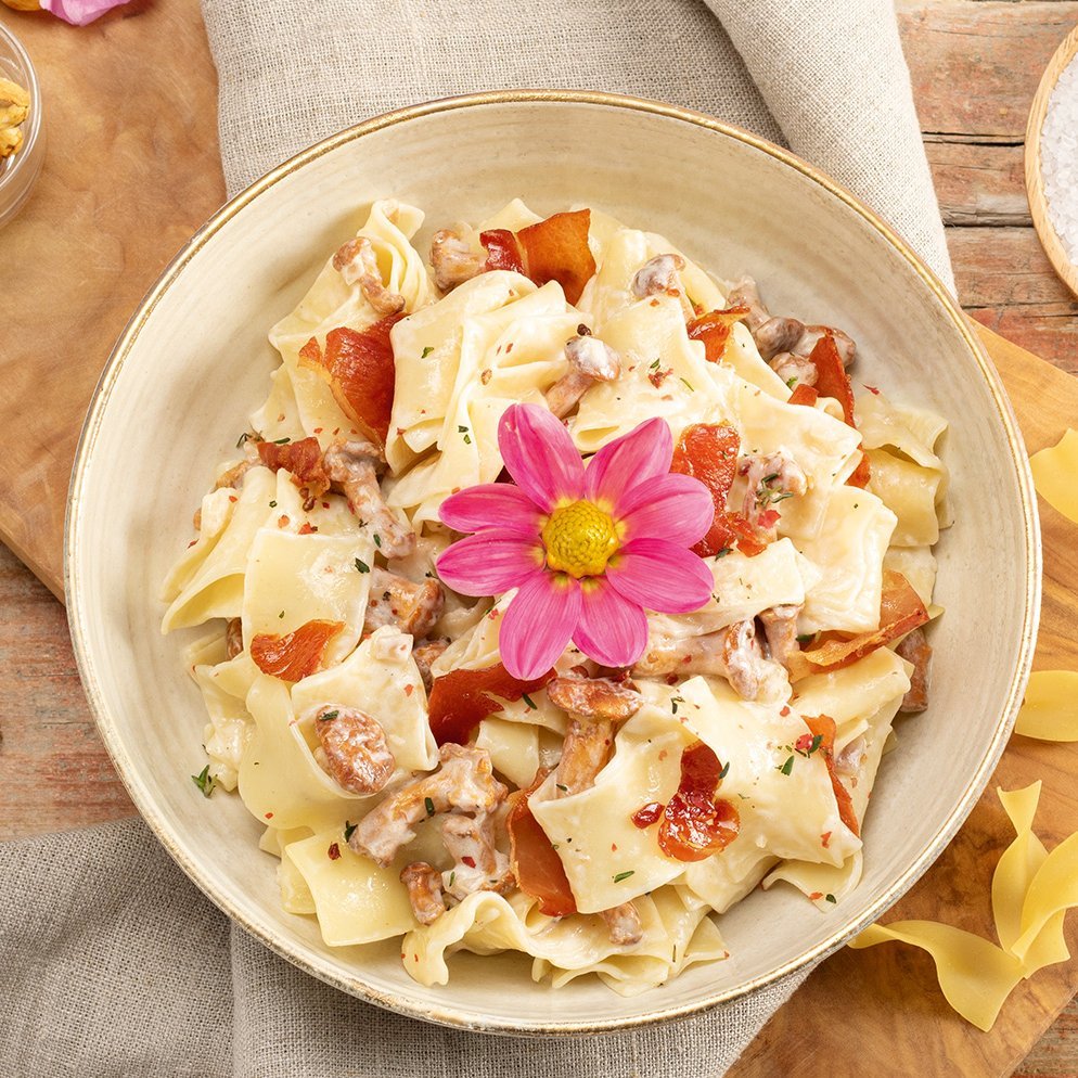 Schüssel mit cremigem Nudelsalat, garniert mit Speckstücken und einer pinken Blume in der Mitte