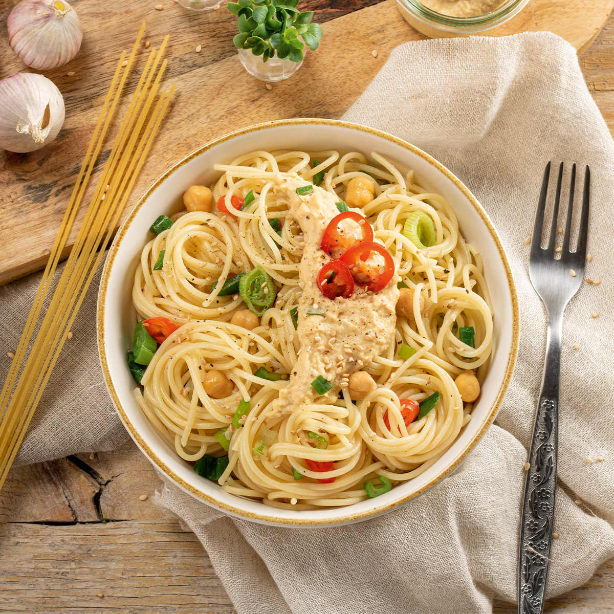  Bowl with spaghetti, chickpeas, cherry tomatoes and herbs on a linen cloth, fork and raw spaghetti next to it