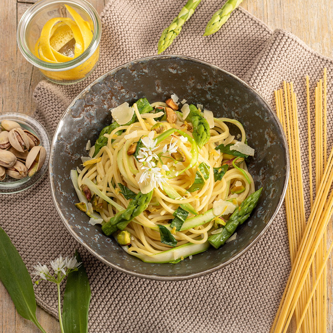 [Translate to Englisch:] Schüssel mit Spaghetti, Spargelstücken und Parmesan auf einem Tisch mit rohem Spargel und ungekochten Spaghetti